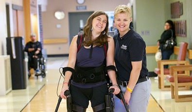 A women uses an exoskeleton to walk down the hospital hall with her therapist.